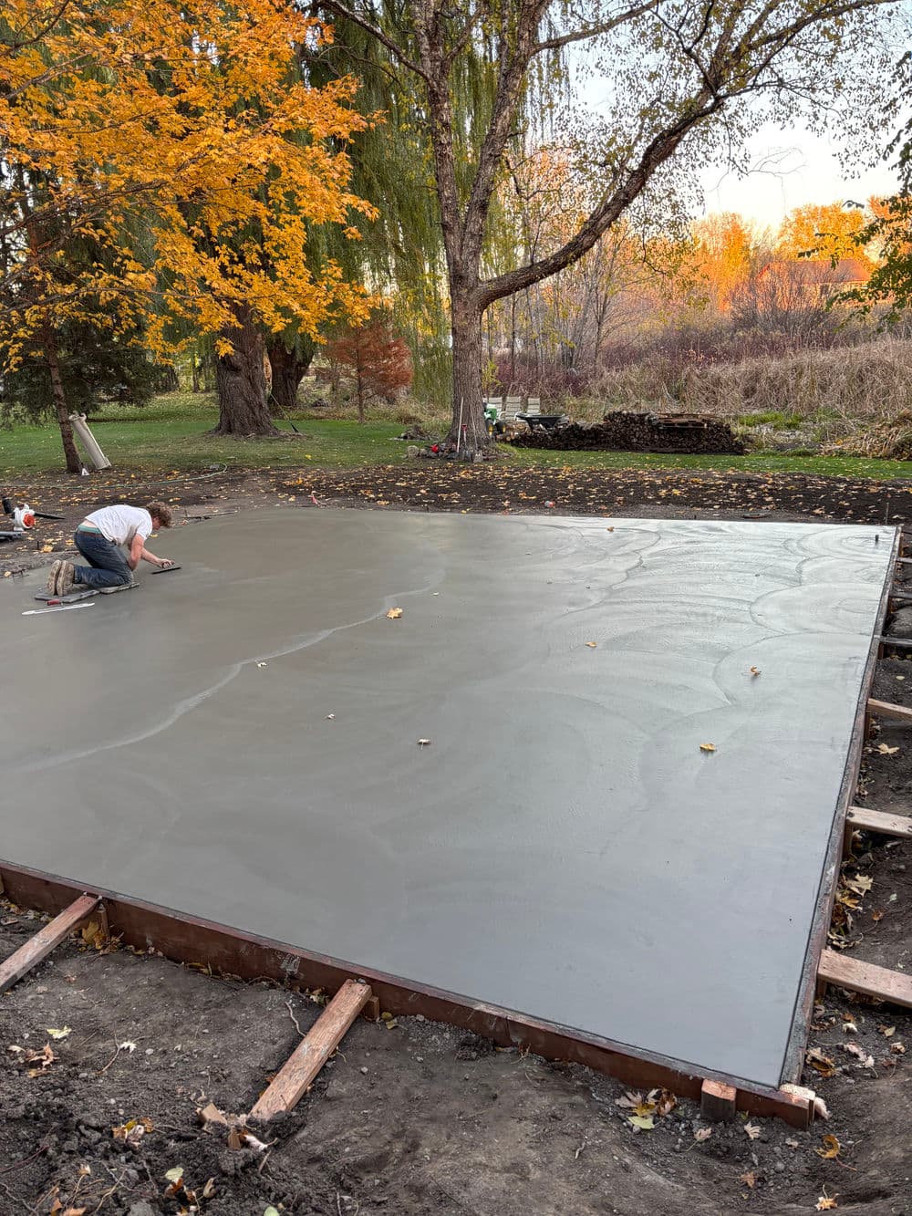 Worker smoothing freshly poured concrete slab in autumn landscape with colorful trees.