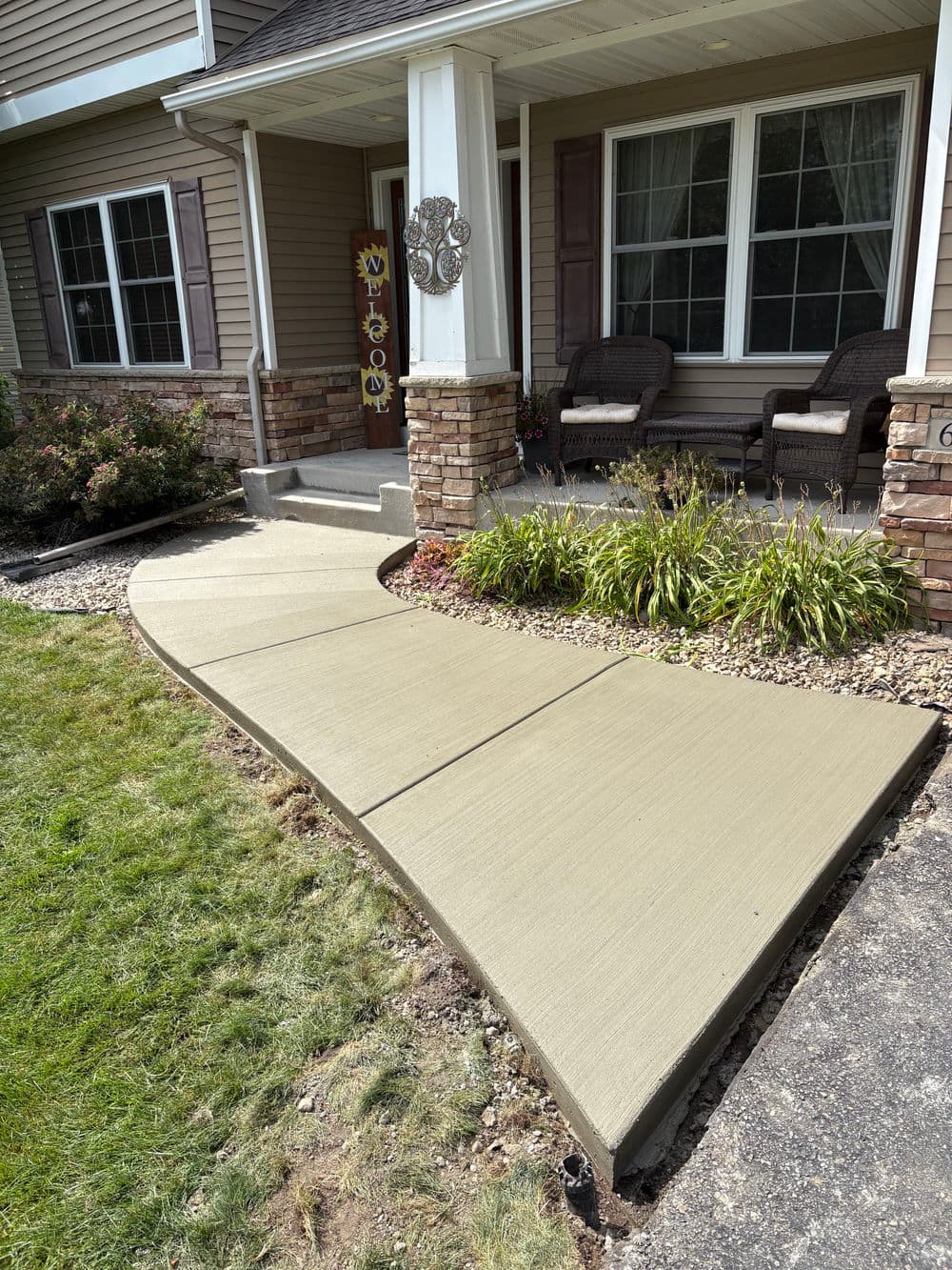 Curved concrete walkway leading to a home entrance with plants and porch furniture.