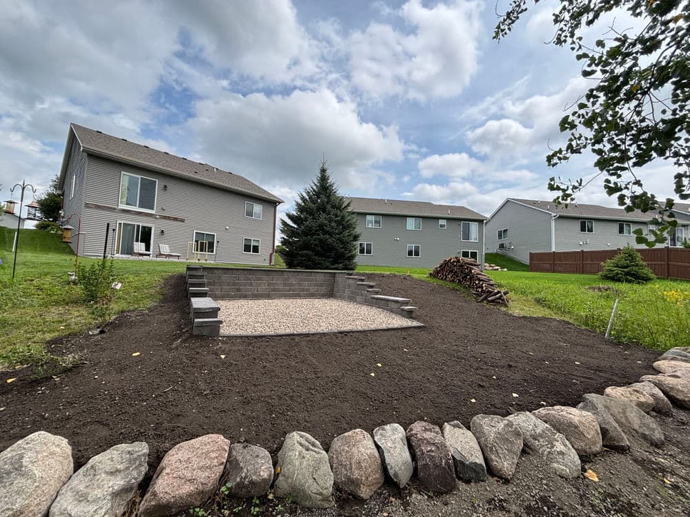 Landscaped backyard featuring a stone patio and pine tree surrounded by homes and a woodpile.