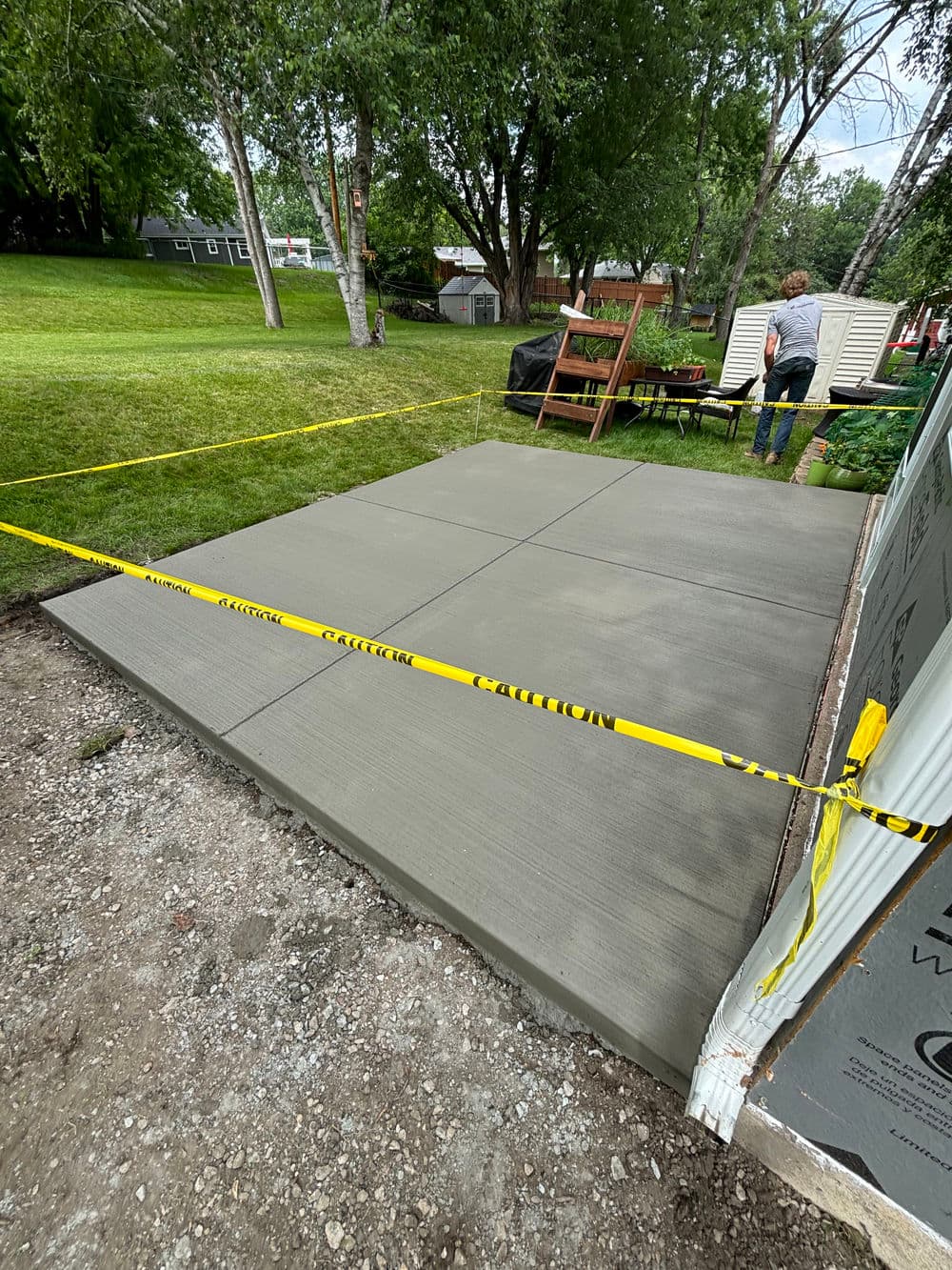 Freshly poured concrete slab in a backyard, surrounded by caution tape and trees.