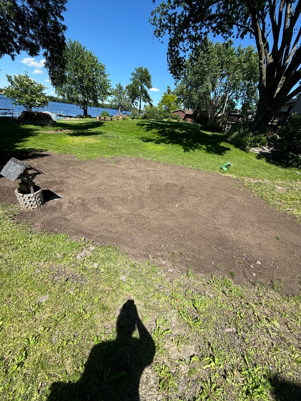 Bare soil area in a sunny park near a lake, surrounded by trees and grass.