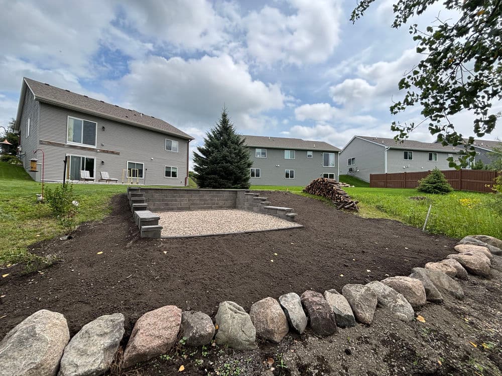 Backyard landscape featuring a gravel area, stone border, and nearby houses under a cloudy sky.