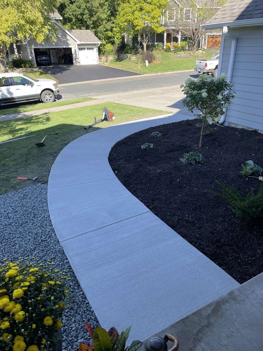 Curved concrete walkway leading to a house, surrounded by landscaped garden and driveway.