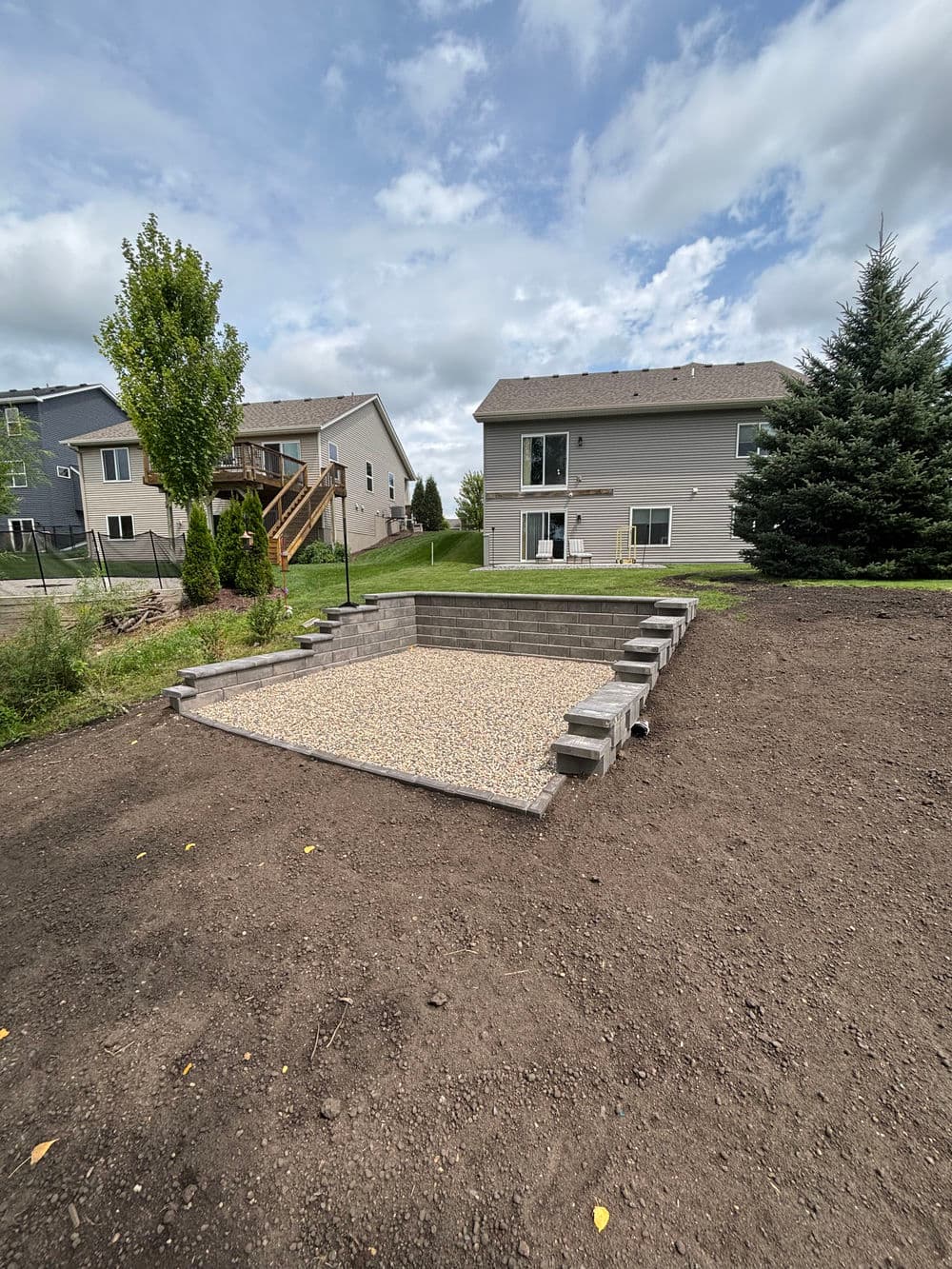Backyard patio area with gravel base and stone steps, surrounded by homes and trees.